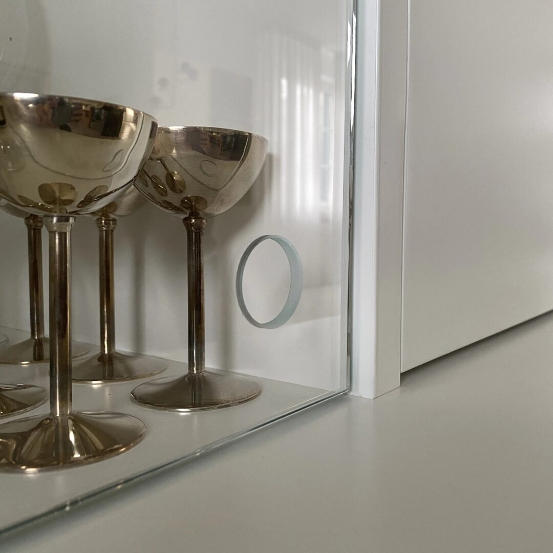 Nahaufnahme einer Glastür mit rundem Lochgriff an einem weißen Schrank gefüllt mit Weingläsern aus Metall