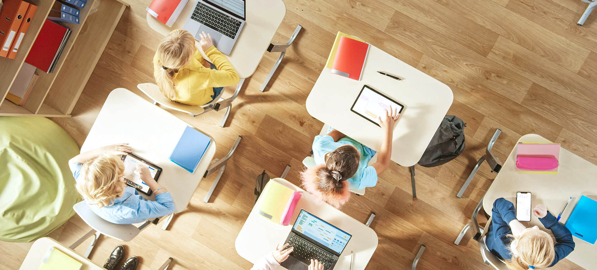 Vogelperspektive auf 5 Kinder, die an quadratischen Einzeltischen mit digitalen Geräten in einem Klassenzimmer lernen